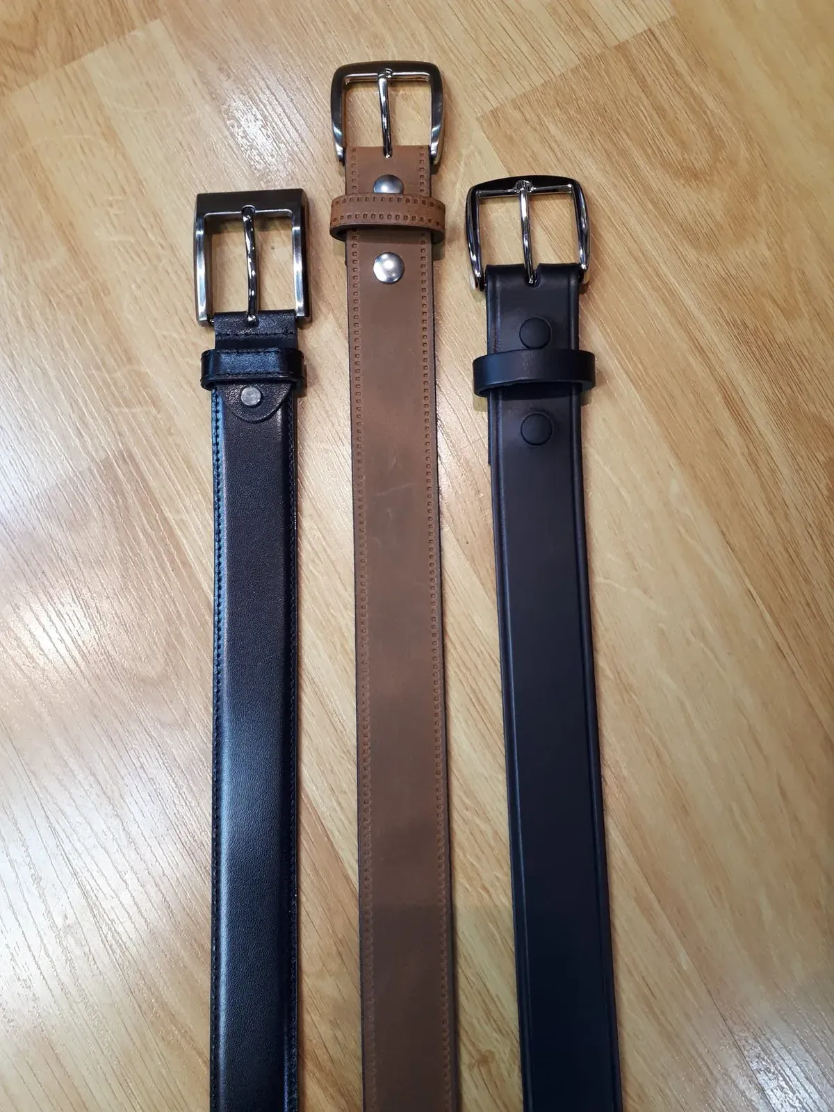 Trois ceintures de cuir alignées — noire, brune piquée, noire à œillets — sur le comptoir.