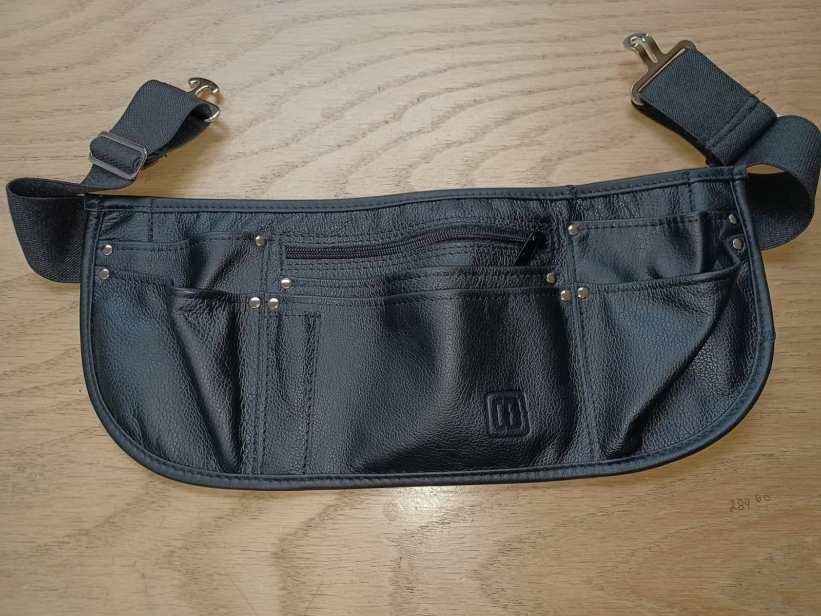 Sacoche de taille en cuir noir à plusieurs poches, sangle réglable, posée sur le comptoir de bois de l’atelier.